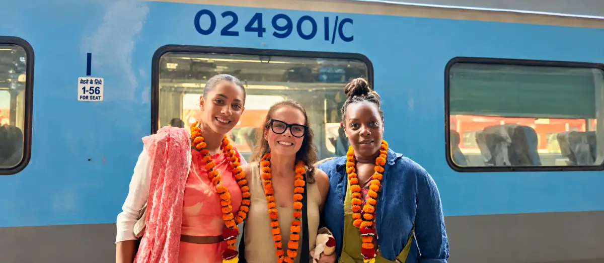 Three female tourists wearing marigold garlands posing beside a train at Agra Cantt Railway Station in India.